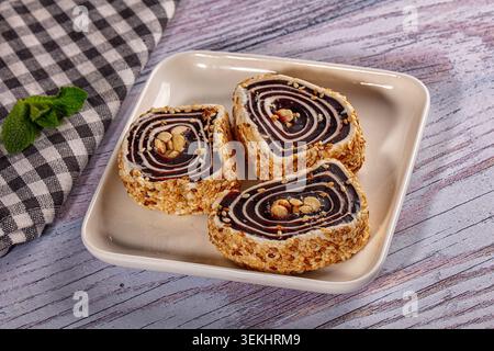 Tre pezzi di dolce arrotolato con ripieno a spirale scuro, noci e semi di sesamo. Servito su un piatto quadrato bianco su un tavolo di legno azzurro con scacchi Foto Stock