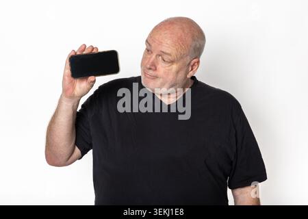 Uomo anziano in maglietta nera che regge il mockup dello smartphone con schermo vuoto e spazio di copia su sfondo bianco Foto Stock