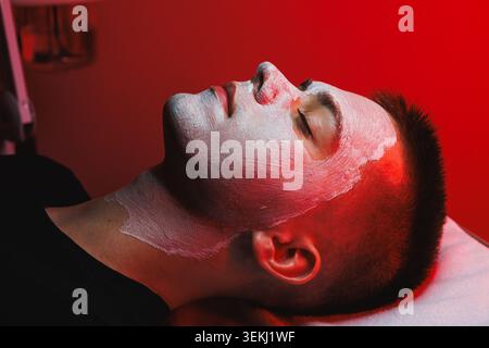 Un cliente riceve una terapia a luce rossa ad un appuntamento cosmetologico. L'individuo è sdraiato su un letto di trattamento con una maschera facciale, circondato da rosso Foto Stock