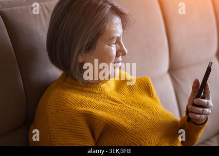 Donna anziana che si rilassa su un divano e utilizza uno smartphone in un accogliente soggiorno con luce a finestra Foto Stock