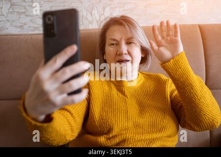 Donna anziana che effettua una videochiamata da un divano e sventola al suo smartphone in un ambiente accogliente Foto Stock
