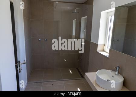 Un bagno contemporaneo con grande cabina doccia piastrellata, un lavandino bianco e una finestra luminosa che fornisce luce naturale Foto Stock
