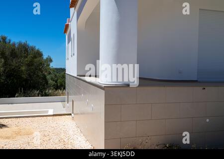 Vista esterna di una casa contemporanea caratterizzata da pareti bianche, una grande colonna e rivestimento in pietra, circondata da una vegetazione vibrante e da un cielo limpido Foto Stock