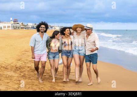 Gruppo di amici sorridenti che camminano a piedi nudi lungo una spiaggia di sabbia soleggiata con le braccia l'una intorno all'altra, godendosi una spensierata vacanza estiva all'oce Foto Stock