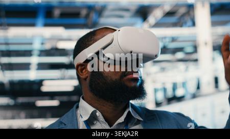 Ritratto di un uomo sorridente nel data center che indossa il visore VR per ottimizzare l'elettronica. Allegro amministratore di sistema di server hub che utilizza la simulazione della realtà virtuale per monitorare le prestazioni del sistema, telecamera B. Foto Stock
