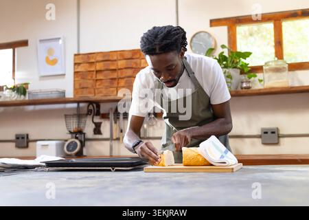 Uomo afro-americano in grembiule verde che affetta la pagnotta di pane su un bancone di legno con coltello Foto Stock
