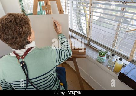 Vista a spalla di una donna matura in un maglione a righe che si avvicina in avanti per dipingere su una tela vuota su un cavalletto, con opere d'arte sul davanzale Foto Stock