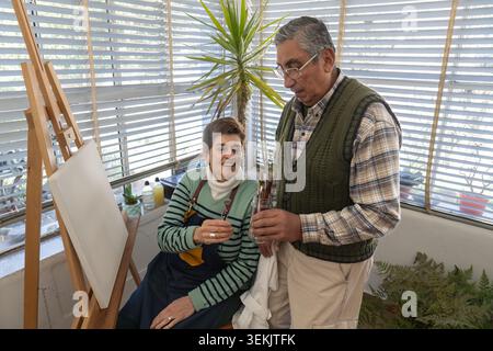 Coppia senior che sceglie i pennelli insieme accanto a una tela vuota in un luminoso studio di arte domestica. Foto Stock