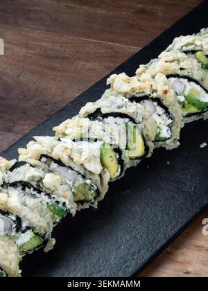 La pastella croccante e dorata copre ogni sushi roll. Avocado e pesce bianco riempiono gli involtini con cura. Il sushi si appoggia su un piatto da portata scuro ed elegante. In legno su Foto Stock