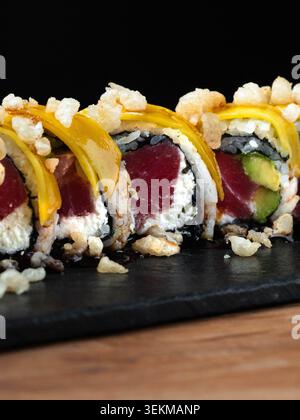 Le fette di pesce dorato adornano i croccanti panini di riso. Punte croccanti e avocado verde aggiungono consistenza. La piastra in ardesia nera poggia su una superficie di legno calda. Sushi artis moderno Foto Stock