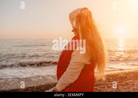 Donna tramonto spiaggia giovane donna bionda in un gilet rosso gode del caldo bagliore del tramonto su una spiaggia di ciottoli in riva al mare Foto Stock