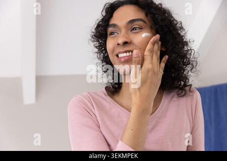Persona non-binaria che applica crema bianca per dare la sfrontatura alla vanità, indossa la parte superiore rosa, anelli e collana Foto Stock