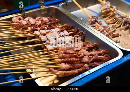 Spiedini freschi di calamari e polpo su vassoi di metallo in vendita al mercato notturno di Temple Street, al chiosco di cibo di strada sotto le calde luci serali di Hong Kong Foto Stock