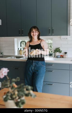 Ritratto di una donna felice che tiene la ciotola in piedi vicino al bancone della cucina a casa Foto Stock