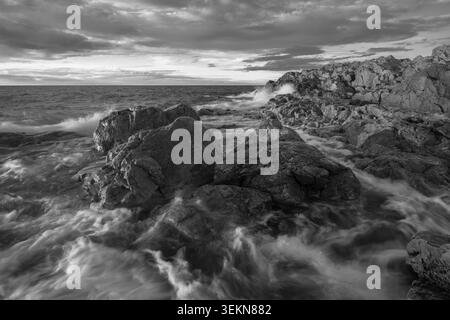 Immagine in bianco e nero di onde che si infrangono su rocce contro il cielo Foto Stock