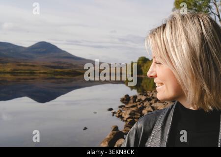 Donna bionda sorridente con gli occhi chiusi vicino al lago nella foresta Foto Stock