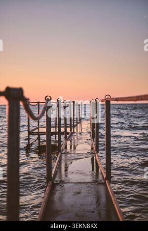 Uno stretto molo in metallo con ringhiere in corda si estende su un'acqua lievemente ondulata verso l'orizzonte, illuminato da caldi colori del tramonto e dalla luce soffusa della sera Foto Stock
