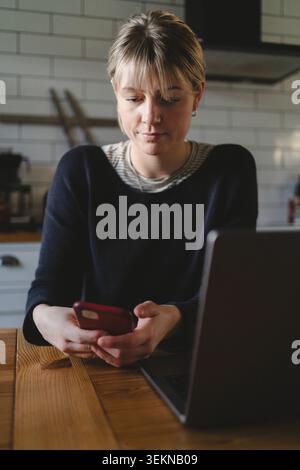 Donna bionda che usa lo smartphone mentre siede con un computer portatile a un tavolo a casa Foto Stock