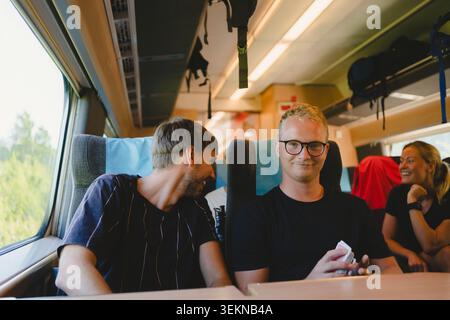 Ritratto di un giovane sorridente che indossa occhiali da vista che viaggia con un amico maschio in treno Foto Stock