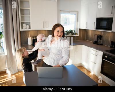 Vista ad angolo alto della ragazza che gioca con il fratello del bambino mentre la madre lavora al laptop a casa Foto Stock