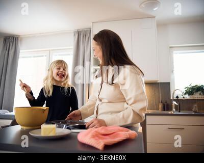 Una bionda giocosa che lecca la pastella dei muffin mentre trascorri il tempo libero con la mamma a casa Foto Stock