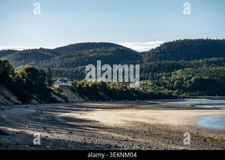 Paesaggi con bassa marea lungo le spiagge di Baie Sainte Catherine, con coste sabbiose e acciottolate, acque basse e calme, rocce sparse e colline boscose sullo sfondo sotto un cielo azzurro Foto Stock