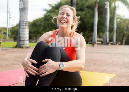Una donna matura e felice con una canotta arancione si siede su un tappetino da yoga in un parco, abbracciandosi le ginocchia e ridendo dopo aver terminato la sua routine di fitness. Foto Stock