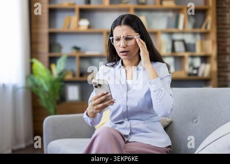 Giovane donna stressata che indossa gli occhiali che prova un'emicrania e tocca il suo tempio, riceve cattive notizie sul suo smartphone, sente ansia, confusione e disagio a casa Foto Stock