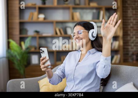 Giovane donna con le cuffie che sorride e saluta lo smartphone durante una rilassante videochiamata sul divano, ascoltando musica e collegandosi online in un accogliente salotto Foto Stock