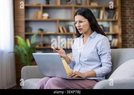 Donna che indossa gli occhiali seduti su un divano e guarda il suo computer portatile con un'espressione confusa e stressata, che ha a che fare con un errore o un problema durante il lavoro a distanza a casa Foto Stock