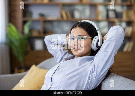 Giovane donna che indossa occhiali trasparenti e cuffie bianche che si rilassa sul divano, ascolta la musica e sorride davanti alla fotocamera in un comodo salotto Foto Stock
