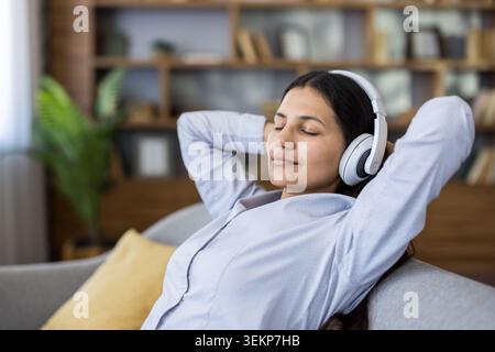 Una giovane donna sdraiata sul divano a casa con gli occhi chiusi, indossa le cuffie e goditi la musica tranquilla per rilassarsi, prendersi cura di sé e rilassarsi in un moderno soggiorno Foto Stock