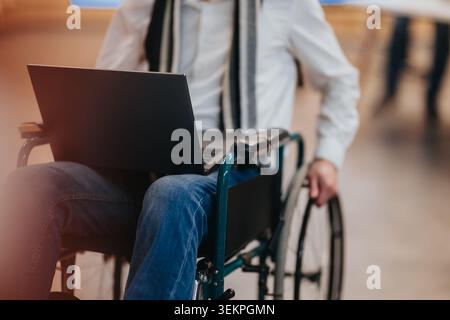 Un uomo d'affari in sedia a rotelle che utilizza un computer portatile nel corridoio dell'ufficio si concentra sul lavoro remoto e sull'accessibilità Foto Stock