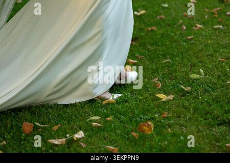 Sposa che cammina sull'erba verde con foglie cadute, indossa un abito da sposa bianco e scarpe Foto Stock