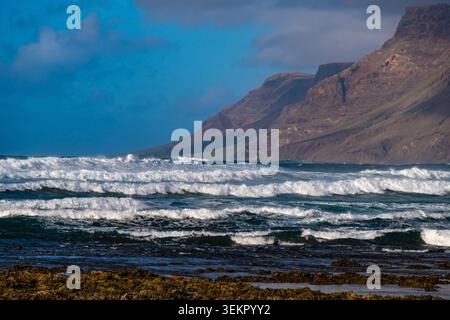 Le onde si infrangono sulla spiaggia di Famara a Lanzarote. La spiaggia sabbiosa incontra l'oceano, mentre le nuvole si radunano sulle scogliere sullo sfondo. Alla gente piace la natura Foto Stock