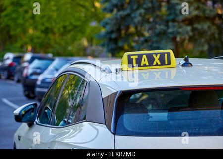 Primo piano di un classico cartello giallo per taxi con scritta nera montato sul tetto di una moderna auto di colore chiaro parcheggiata lungo una strada della città. Foto Stock