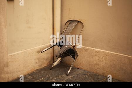 Una singola sedia in metallo usurata che si appoggia in modo imbarazzante in un angolo dell'edificio, fissata saldamente con una catena a un vecchio tubo di drenaggio verticale. Foto Stock
