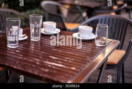 Primo piano di tazze da caffè bianche vuote con piattini e bicchieri d'acqua trasparenti lasciati su un tavolo di legno scuro in una terrazza all'aperto. Foto Stock