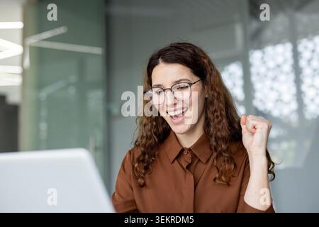 Giovane donna allegra con gli occhiali che sorride e alza il pugno in un ufficio moderno mentre guarda un notebook, suggerendo risultati, buone notizie o successo Foto Stock