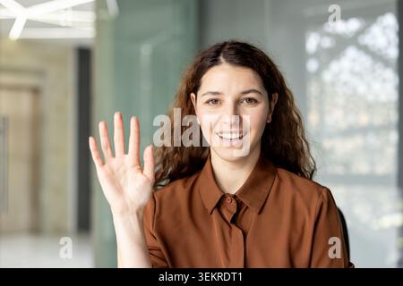 Giovane donna amichevole con lunghi capelli ricci sorridente e alzata la mano nel saluto all'interno di un luminoso spazio ufficio moderno con vetro sfocato e finestre nella Foto Stock