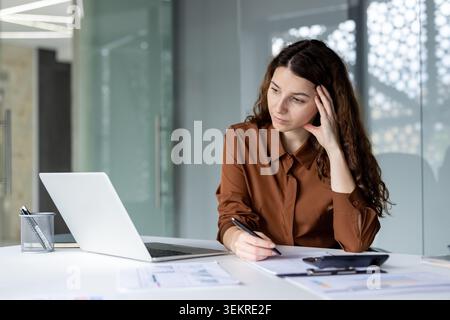 Una giovane donna siede a una scrivania con un laptop e documenti in un ufficio luminoso e moderno, appoggiando la testa su una mano mentre scrive e sembra penosa Foto Stock