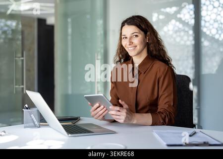 Giovane donna seduta a una scrivania in un ufficio luminoso e moderno, sorridente davanti alla fotocamera mentre tiene un tablet accanto a un laptop aperto e documenti, con pareti in vetro Foto Stock
