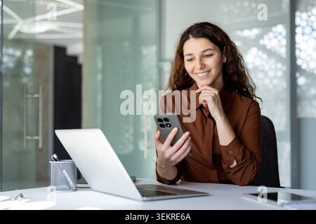 Giovane donna seduta a una scrivania moderna, sorridente mentre guarda il suo smartphone accanto a un laptop aperto in un luminoso spazio di lavoro con pareti in vetro Foto Stock