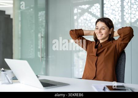 Giovane donna sorridente seduta a una scrivania con le mani dietro la testa in un ufficio luminoso e moderno, con un laptop in primo piano e pareti di vetro dietro di lei Foto Stock