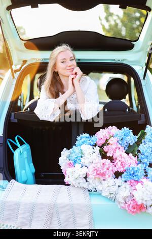 Allegra viaggiatrice che posa in un bagagliaio compatto con fiori e accessori da picnic durante un weekend rilassante Foto Stock