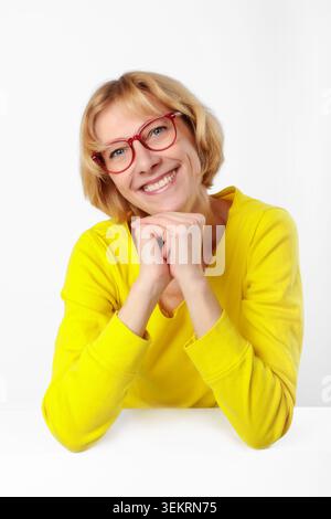 Ritratto in studio di una donna amichevole e premurosa con occhiali che indossa un maglione giallo brillante, seduta a tavola su sfondo chiaro, apertura, fiducia Foto Stock