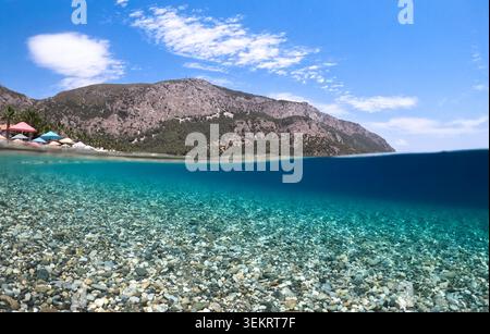 Sotto la spaccata di una spiaggia di ciottoli e di un mare turchese con acqua cristallina, montagne costiere rocciose e cielo blu: Viaggi estivi, resort e natura Foto Stock