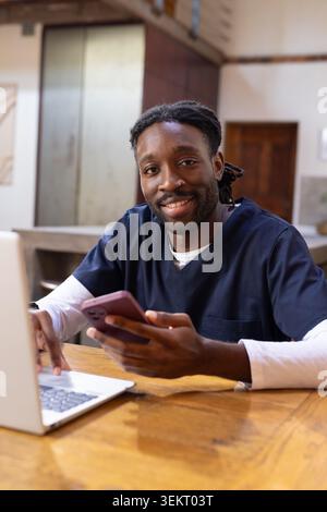 Uomo afro-americano che indossa scrub della marina seduto al tavolo della cucina tenendo il telefono rosa, usando un computer portatile Foto Stock