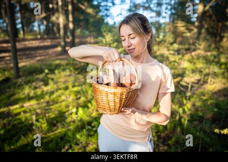 Una donna sorridente e allegra che raccoglie funghi tenendo in mano un cesto di vimini pieno di funghi porcini commestibili Foto Stock