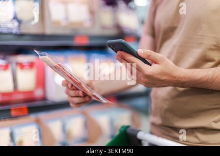 Uomo che confronta il cibo confezionato con lo smartphone nella corsia del supermercato, cliente che controlla le informazioni sul prodotto Foto Stock